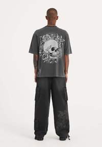 Camiseta gris de gran tamaño con un gran gráfico de calavera y rosa en la parte de atrás. Combinada con pantalones cargo negros holgados con bolsillos laterales y bordado.