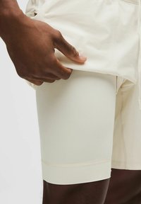 Mano levantando unos pantalones cortos beige para revelar unos pantalones cortos de compresión ajustados blancos debajo, sobre un muslo de piel oscura con fondo blanco.