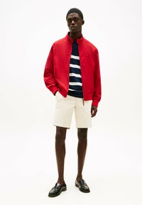Rød bomberjakke over en marineblå stripete skjorte, kombinert med kremfargede shorts og svarte loafers. Glatt stoff med en sporty silhuett.