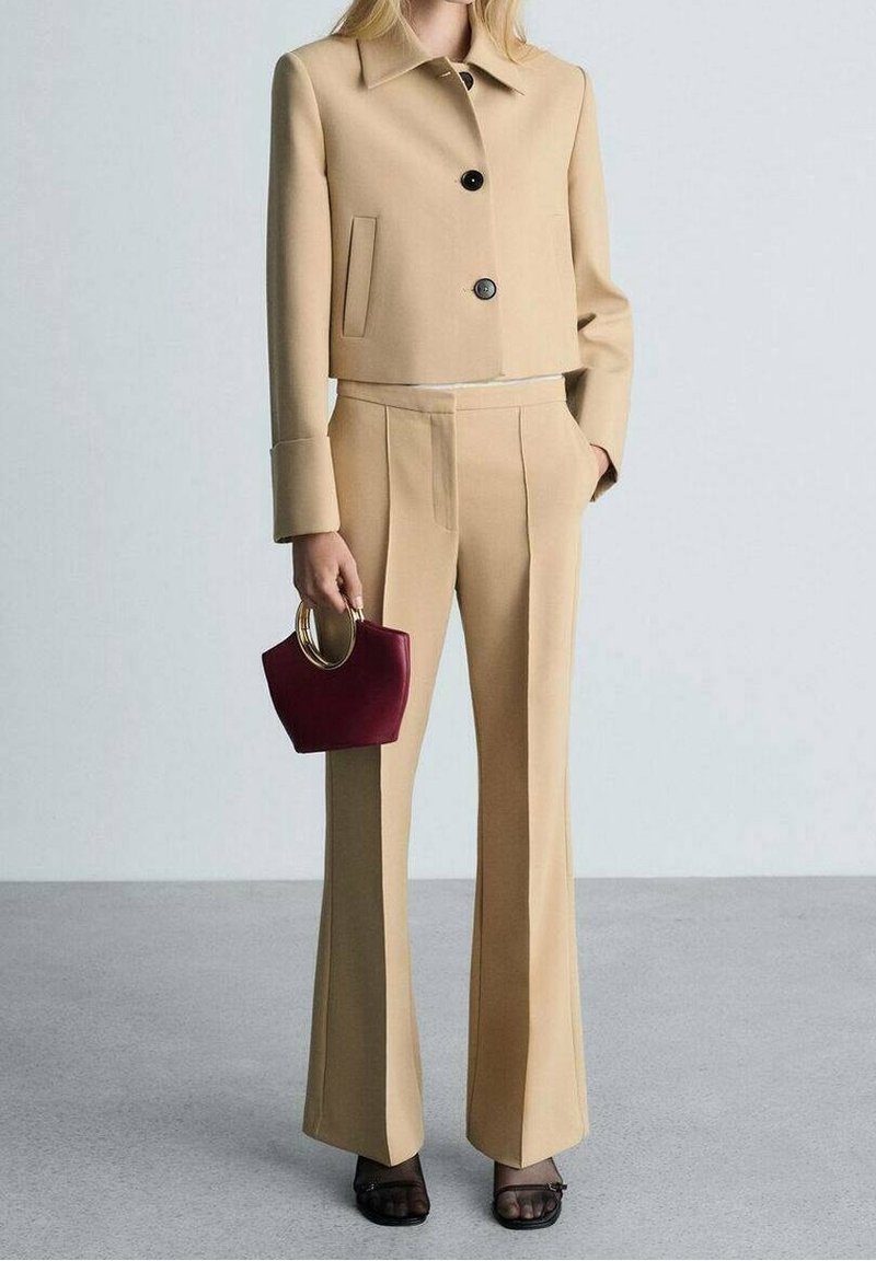 Femme portant une veste courte beige cintrée et un pantalon taille haute assorti, tenant un petit sac à main bordeaux avec des anses dorées, chaussures noires à bout ouvert.