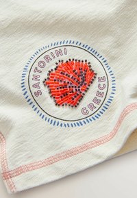 Bílá látka s kruhovým vzorem obsahujícím červenou a černou korálkovou lasturu uprostřed, obklopenou textem "Santorini Řecko" a modrými čárkami.