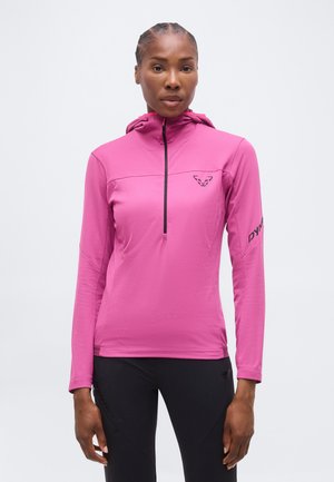 Kobieta w długim, jaskrawo różowym sportowym topie z kapturem i zamkiem z przodu oraz czarnych spodniach, stojąca na jednolitym tle.