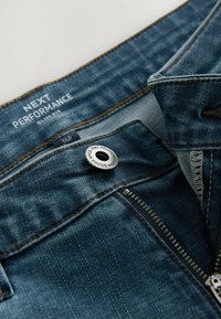 Jean en denim bleu moyen avec une braguette zippée, des coutures marron et un bouton argenté. L'étiquette indique "NEXT PERFORMANCE SLIM FIT" et la taille 34R.