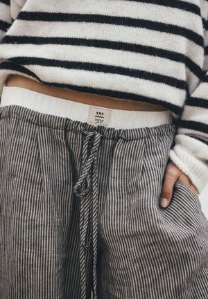 Sweter w paski i wysokie spodnie w paski z sznurkiem, ręka w kieszeni, widoczna metka ubrania SRF nad paskiem spodni.