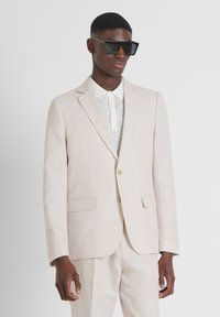 Costume en lin beige clair avec une fermeture à un bouton, des revers en elle, et des poches latérales, associé à une chemise légère texturée et des lunettes de soleil foncées.