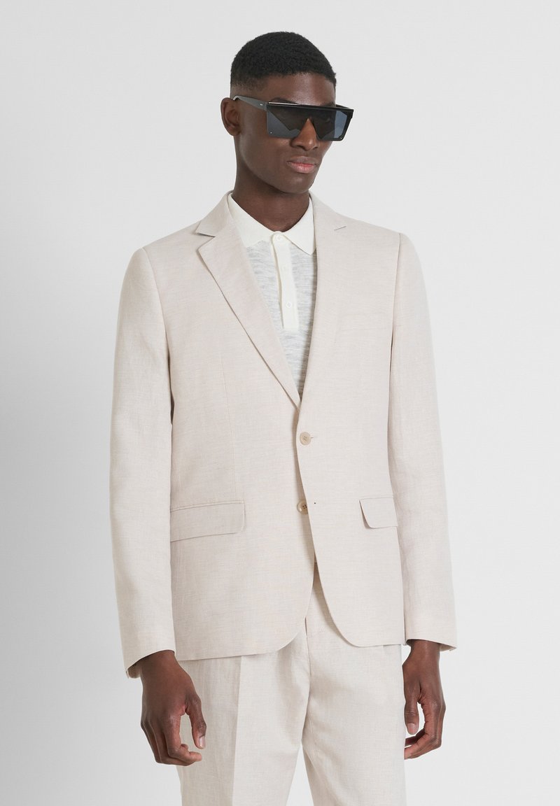 Costume en lin beige clair avec une fermeture à un bouton, des revers en elle, et des poches latérales, associé à une chemise légère texturée et des lunettes de soleil foncées.