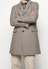 Cappotto in lana grigia chiara con rever a punta, due tasche frontali e chiusura a un bottone. Indossato sopra una camicia a quadri e pantaloni beige.