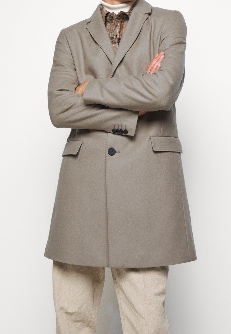 Cappotto in lana grigia chiara con rever a punta, due tasche frontali e chiusura a un bottone. Indossato sopra una camicia a quadri e pantaloni beige.