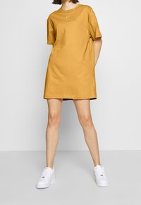 Żółta bawełniana sukienka typu t-shirt z krótkimi rękawami i okrągłym dekoltem. Na piersi znajduje się małe białe logo Nike. Noszona z białymi trampkami.