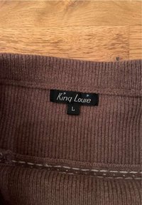 Pull en tricot côtelé marron avec une étiquette noire portant la mention "King Louie" et la taille "L", présentant un détail de couture blanche décorative.
