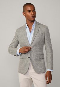 Blazer grigio strutturato con design aderente, due tasche frontali, chiusura a un solo bottone e motivo sottile, indossato sopra una camicia azzurro chiaro.