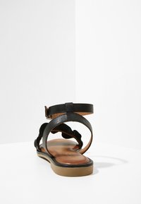 Czarna skórzana sandał z płaską podeszwą, z pojedynczym paskiem na kostkę i falistymi detalami wzdłuż boków. Widoczna beżowa wkładka.