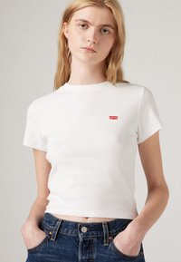 Tricou alb cu maneci scurte, cu dungi, având un mic logo roșu Levi's pe pieptul stâng, purtat cu blugi din denim albastru cu talie înaltă.
