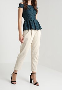 Haut peplum bleu sarcelle avec un design épaules dénudées et un motif texturé, associé à un pantalon en velours côtelé crème et des talons noirs à bride cheville.