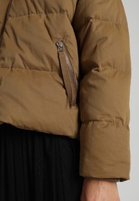 Manche de veste matelassée marron avec poche zippée et languette en tissu marron, portée sur une jupe plissée noire.