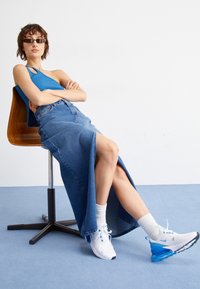 Woman wearing blue one-shoulder top, long denim skirt with slit, white socks, and white-blue sneakers sitting on a wooden swivel chair against plain background.