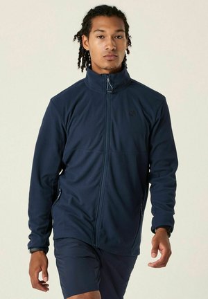 Young man with braided hair wearing a zipped navy fleece jacket and dark shorts walking forward against a plain background.
