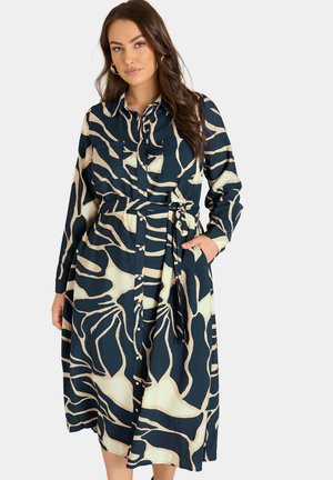 Femme portant une robe à manches longues, boutonnée, à motif noir et beige avec une ceinture à la taille, posant avec une main dans une poche sur un fond uni.