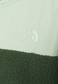 Kurtka polarowa z jasnozieloną górną częścią i ciemnozieloną, teksturowaną dolną częścią z polaru. Zawiera haftowane logo.