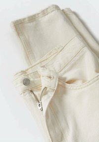 Pantalons en denim de couleur crème avec une fermeture éclair en métal, un bouton de fermeture, des poches avant et des coutures décoratives sur les coutures et la taille.