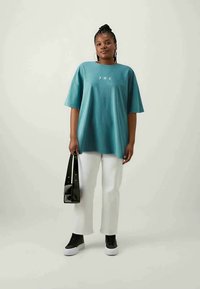 Camiseta azul verdosa holgada con mangas cortas, que presenta un gráfico celeste en blanco. Combinada con pantalones blancos y zapatillas negras. Bolsa negra.