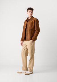 Brown suede jacket with shirt collar, two front pockets, and a tan knit sweater. Light beige trousers and white sneakers with gum soles.