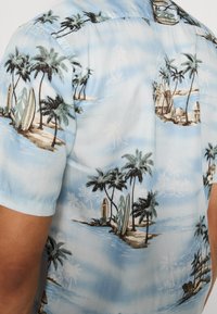 Chemise à manches courtes en tissu bleu clair avec des motifs d'îles tropicales, des palmiers, des planches de surf et des huttes dans un design illustré.