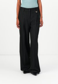 Pantalons noirs à rayures verticales avec une coupe décontractée, taille haute et un accent doré sur la poche. Tissu lisse avec une finition soignée.