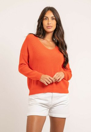 Femme aux longs cheveux foncés portant un pull tricoté orange à manches longues et un short blanc avec ourlet roulé, debout devant un fond uni.