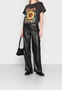 Camiseta negra gráfica con diseño de girasol y texto retro, combinada con pantalones de cuero negro de pierna ancha. La modelo sostiene un bolso negro y lleva botas de plataforma.