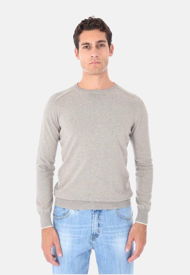 Uomo con capelli corti e ricci, che indossa un maglione grigio chiaro a maniche lunghe e jeans azzurri, in piedi davanti a uno sfondo bianco semplice.