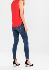 Top rojo sin mangas, jeans ajustados azules y tacones negros con lunares blancos. Enfoque en líneas limpias y ajuste casual.
