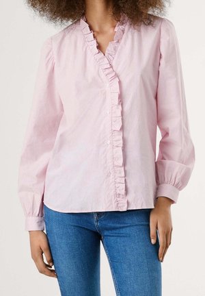 Młoda osoba ubrana w jasnoróżową bluzkę z długim rękawem, ozdobioną falbanką, oraz niebieskie jeansy, stojąca na jednolitym tle.