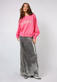 Růžová mikina s nápisem "LOVE" v kombinaci s dlouhou stříbrnou flitrovou sukní. Outfit je charakterizován hladkými texturami a výrazným kovovým leskem.