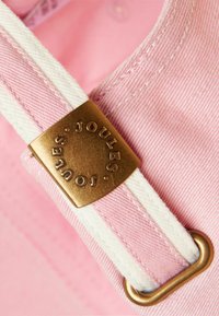 Primer plano de tela rosa con una correa blanca pasada por una hebilla de latón grabada con la palabra "JOULES".