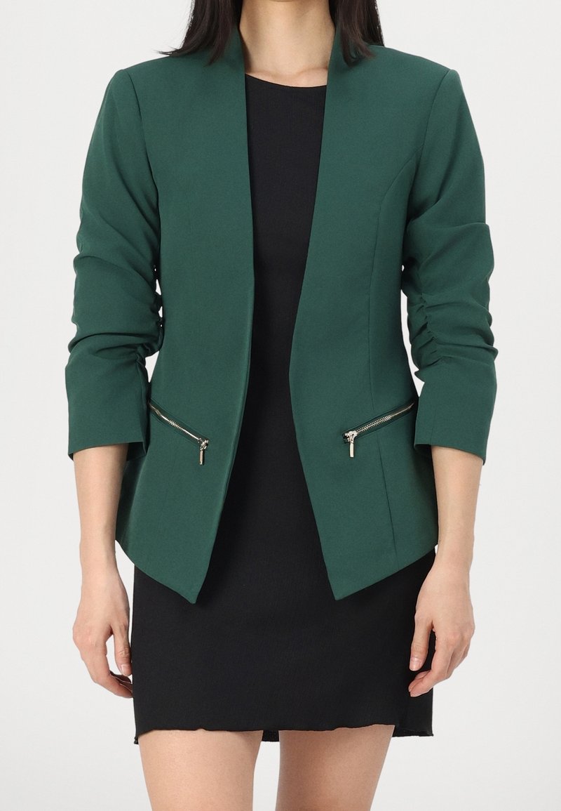 Zelený blazer se strukturovaným střihem, dlouhými rukávy a kapsami na zip. Vyroben z hladkého materiálu, s pevným střihem a otevřeným předním dílem.