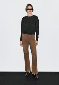 Junge Frau, die eine schwarze, geknöpfte Strickjacke, braune Wildlederhosen, schwarze Loafers und dunkle Sonnenbrillen trägt, steht vor einem schlichten hellen Hintergrund.