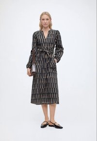 Γυναίκα που φοράει μαύρο και μπεζ φόρεμα με μοτίβο, μεσαίου μήκους και μακριά μανίκια, με δεμένη μέση, κρατώντας καφέ τσάντα ώμου και μαύρες μπαλαρίνες.