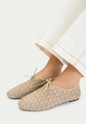 Scarpe piatte beige intrecciate con piccoli fiocchi indossate con pantaloni bianchi a gamba larga, mostrate ai piedi su uno sfondo semplice.