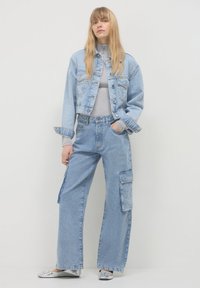 Donna con lunghi capelli biondi che indossa un dolcevita grigio chiaro, una giacca di jeans cropped, pantaloni cargo in denim a gamba larga e ballerine argentate.