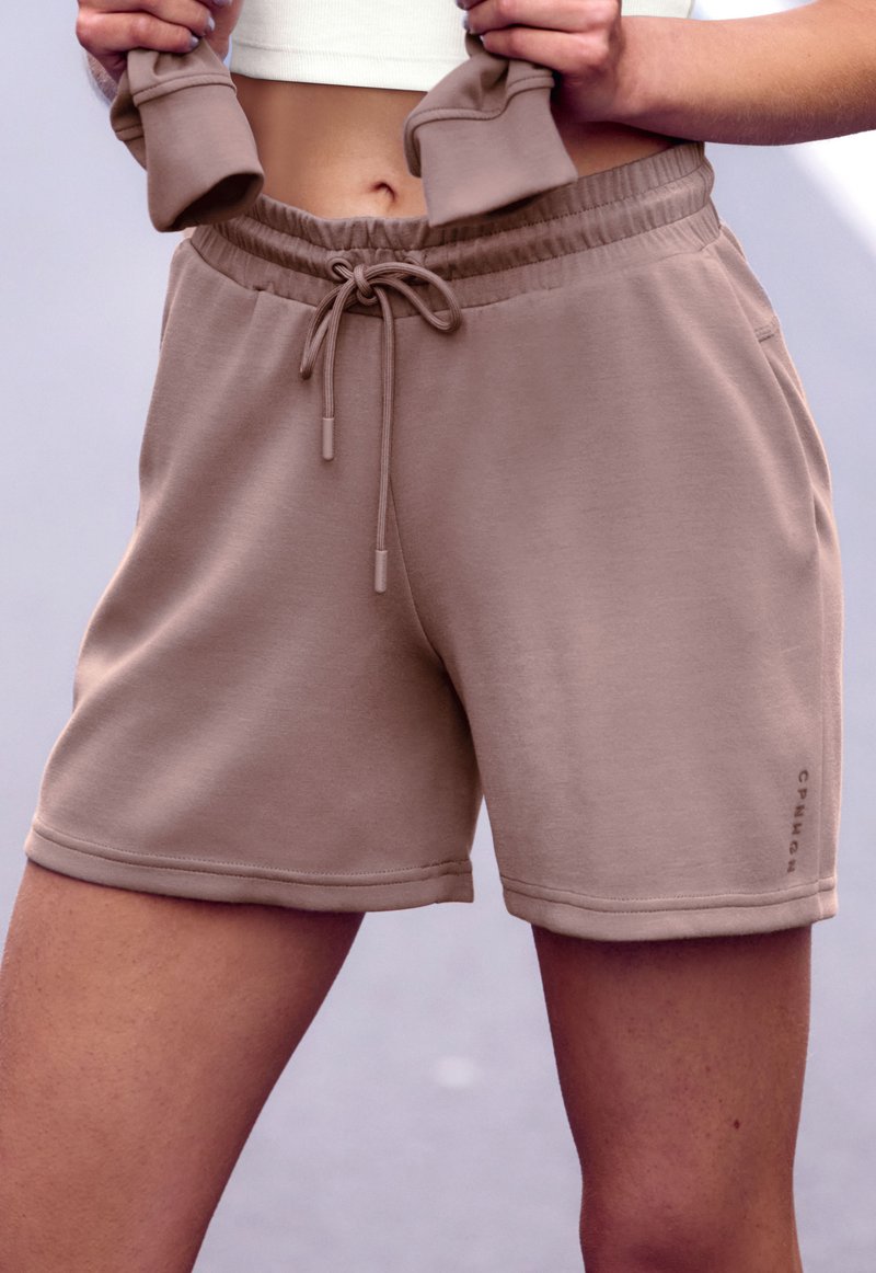 Beige Sportshorts aus weichem Stoff, mit elastischem Bund und Kordelzug sowie seitlichem Logodetail. Glatte Textur und lockere Passform.