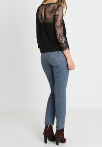 Top nero con maniche e schiena in pizzo floreale trasparente, abbinato a jeans in denim blu e stivaletti con tacco quadrato color bordeaux.