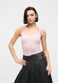 Γυναίκα που φοράει αμάνικο ανοιχτό ροζ τοπ με δαντέλα και μαύρη πλισέ φούστα ψηλόμεση με διπλές αγκράφες.