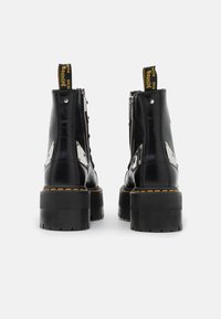 Botas de tobillo de charol negro con tacones gruesos, acentos de herrajes plateados y costuras amarillas. Cierre de cremallera en el lado.