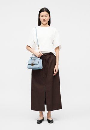 Γυναίκα που φοράει λευκό μπλουζάκι με κοντά μανίκια, σκούρα καφέ μίντι φούστα με σκίσιμο μπροστά, μαύρες μπαλαρίνες, κρατώντας ανοιχτό μπλε τσάντα με αλυσίδα στον ώμο.