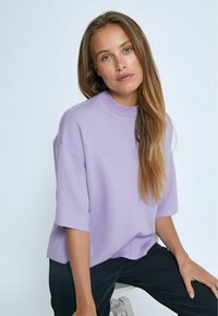 Maglione corto a maniche corte di colore violaceo chiaro con un colletto alto, realizzato in un tessuto morbido. Caratterizzato da una vestibilità comoda e da una texture liscia. Indossato con pantaloni neri.