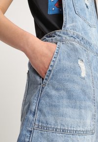 Ljust blå denimoverall med slitna detaljer. Har ett framficka och justerbara axelband. Materialet ser mjukt ut med en avslappnad passform.