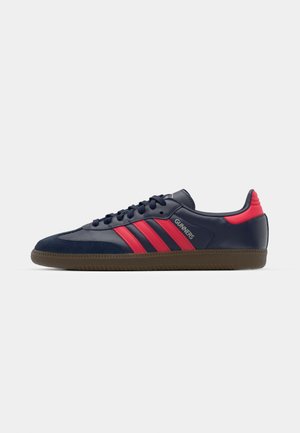 Mørkeblå sneakers med røde striber, gummisål og snørelukning; har en glat syntetisk overflade og et logo på siden.