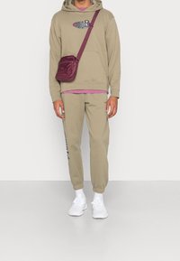 Béžová mikina s logem, navrstvená přes růžovou košili; odpovídající béžové tepláky; malá bordó crossbody taška; bílé tenisky.