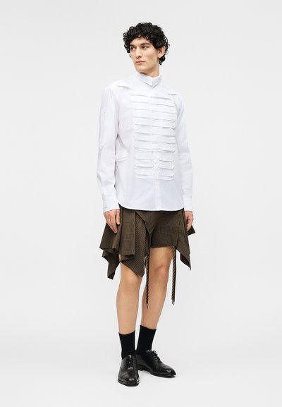 Młoda osoba o kręconych włosach ubrana w białą koszulę zapinaną na guziki z poziomymi plisami oraz asymetryczną brązową spódnicę i czarne buty.